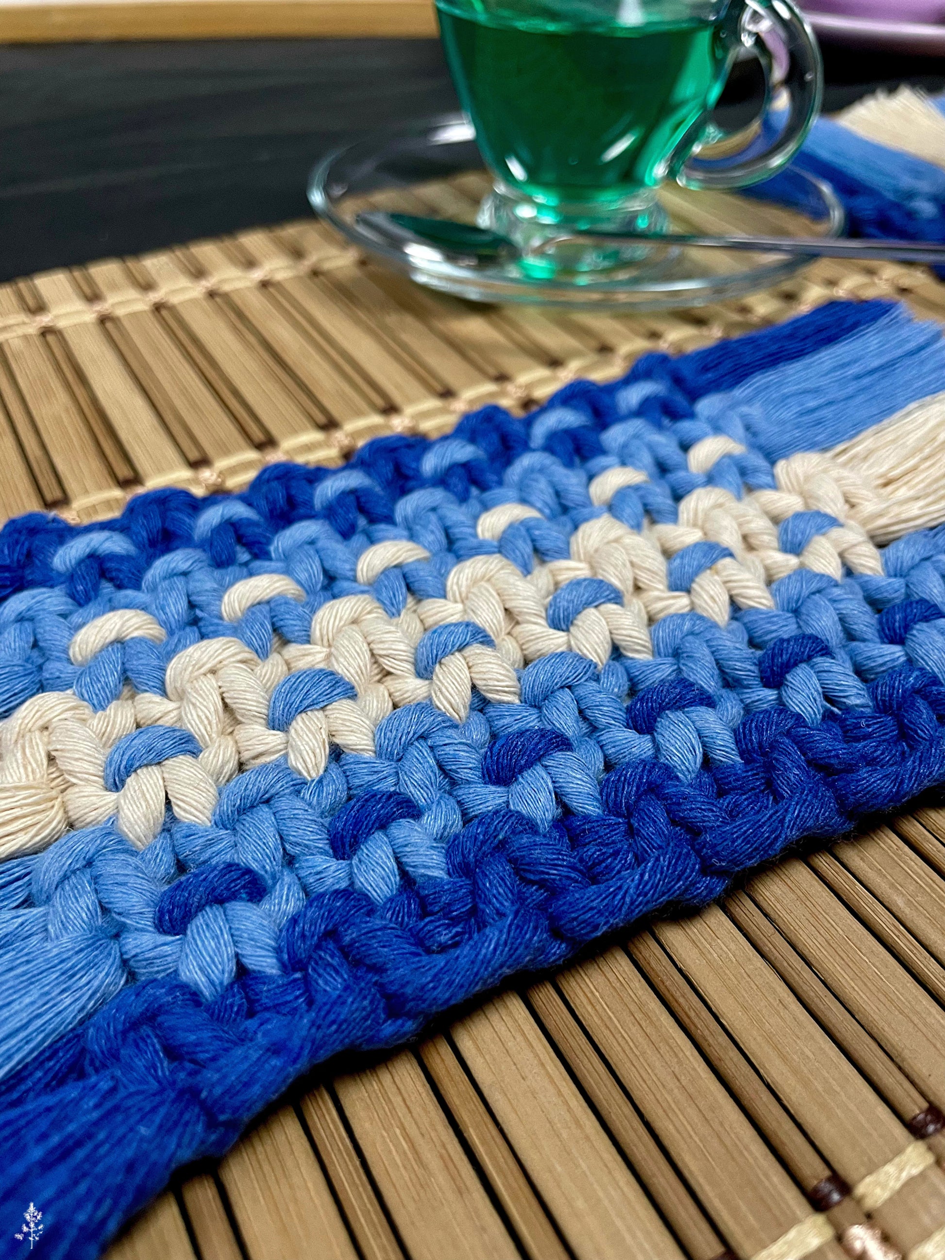 Blue and white macrame coaster on a wooden surface with a glass of green liquid in the background.