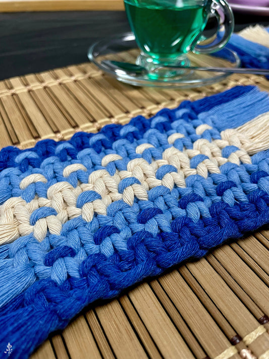 Blue and white macrame coaster on a wooden surface with a glass of green liquid in the background.