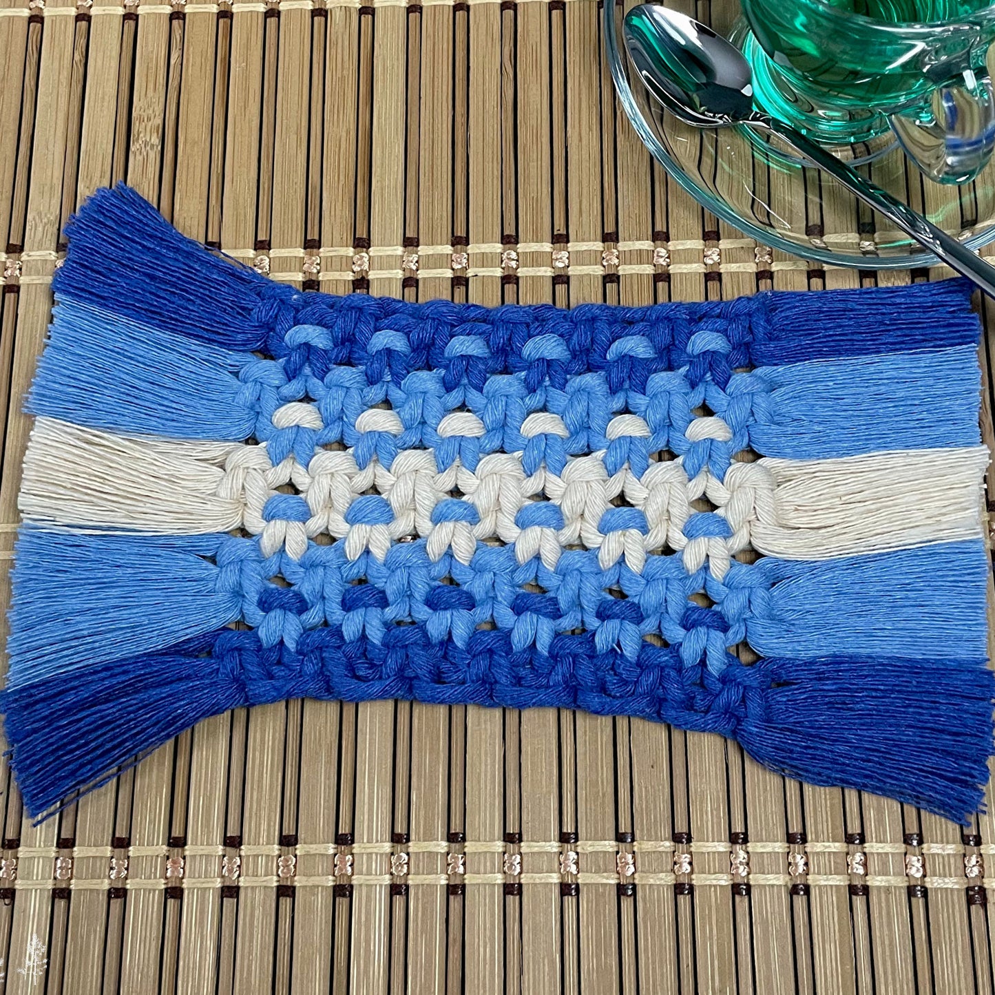 Blue and white macrame coaster on a wooden surface with a glass and spoon.