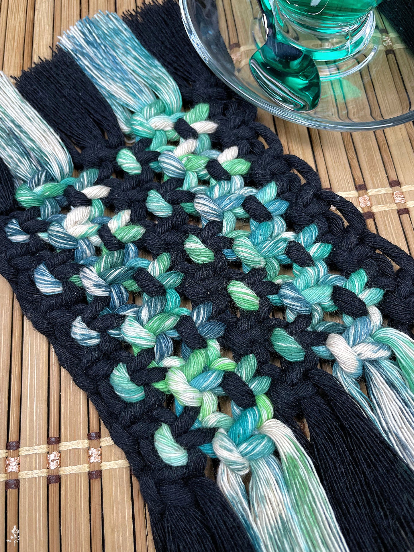 Macrame coaster with black and tie dyed green rope on a wooden surface.