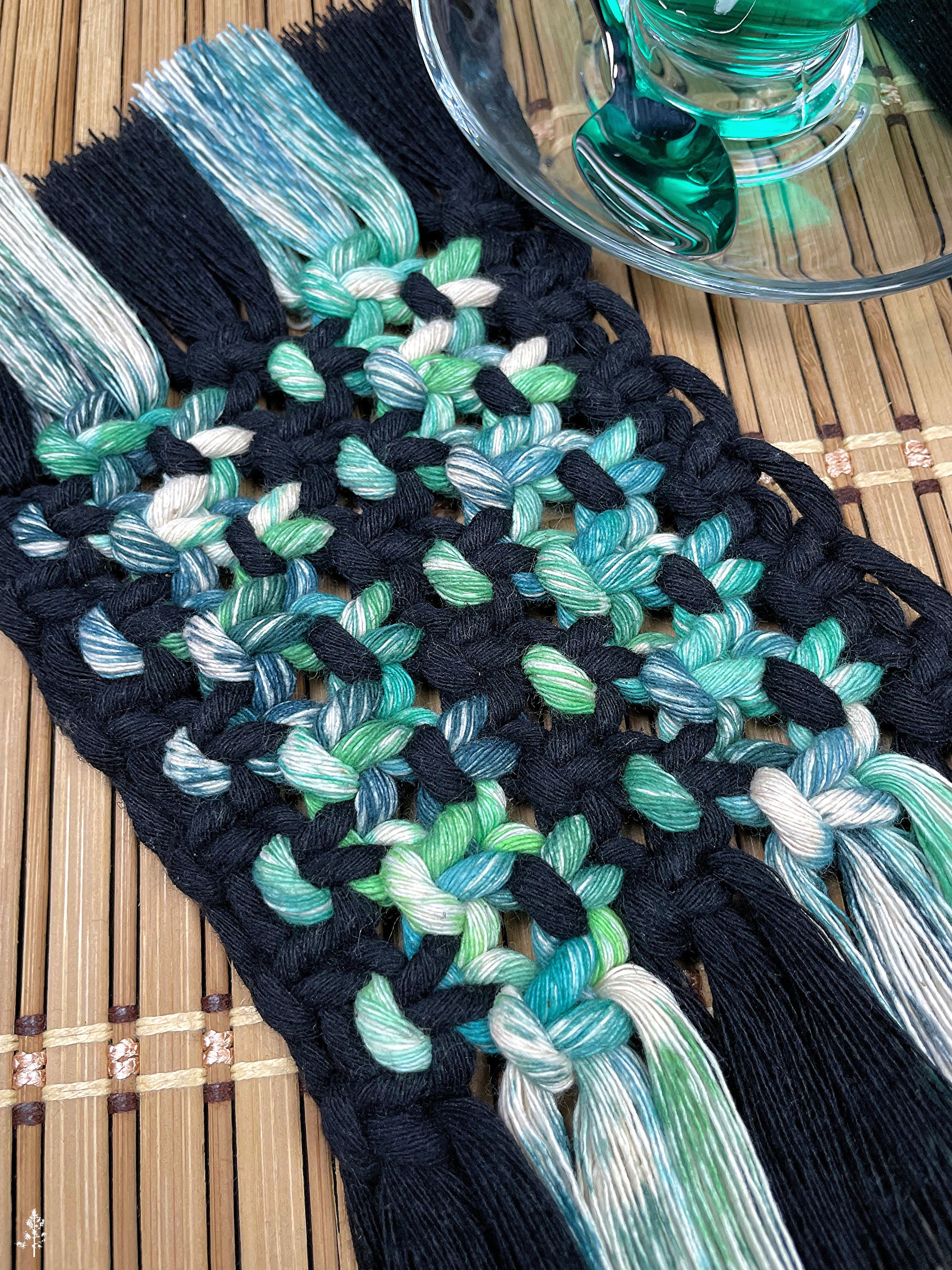 Macrame coaster with black and tie dyed green rope on a wooden surface.