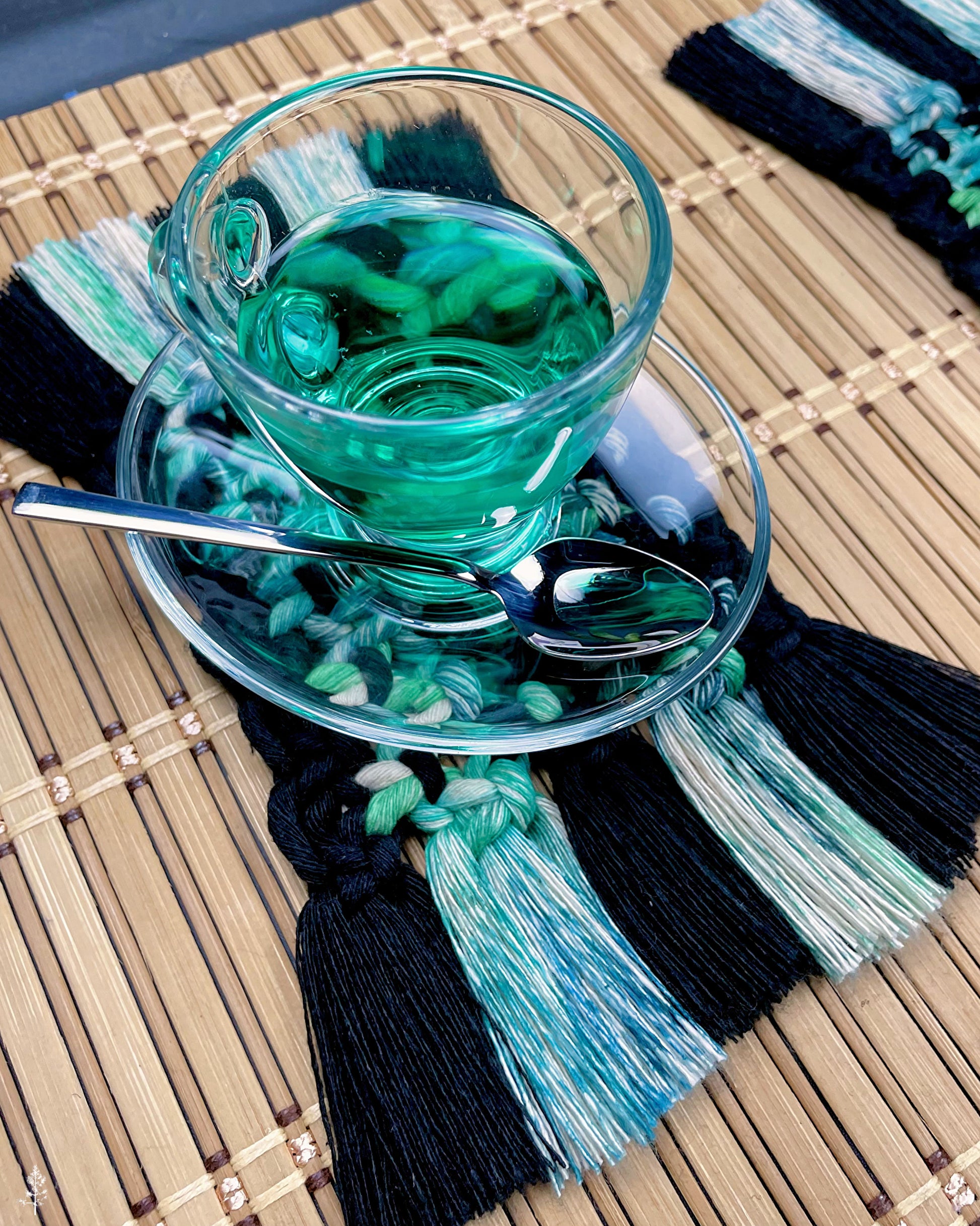 Tea glass, saucer, and spoon resting one a black and tie dye macrame coaster