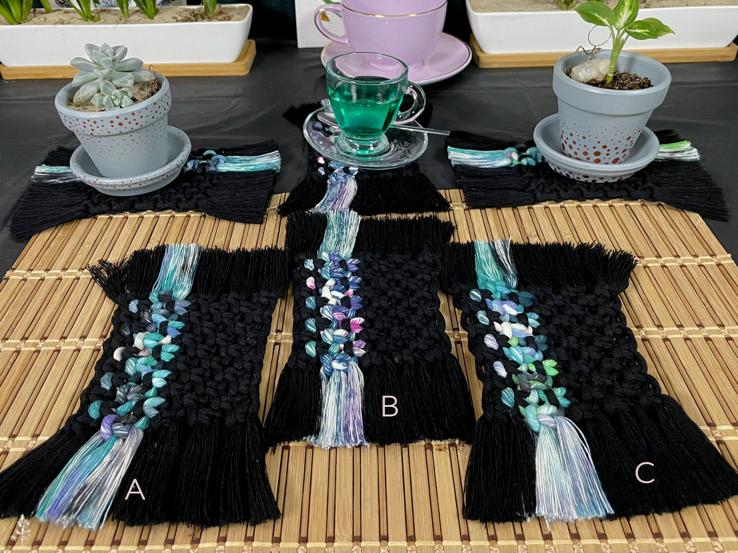 Black macrame coasters with colorful tassels on a wooden table with plants and tea set.