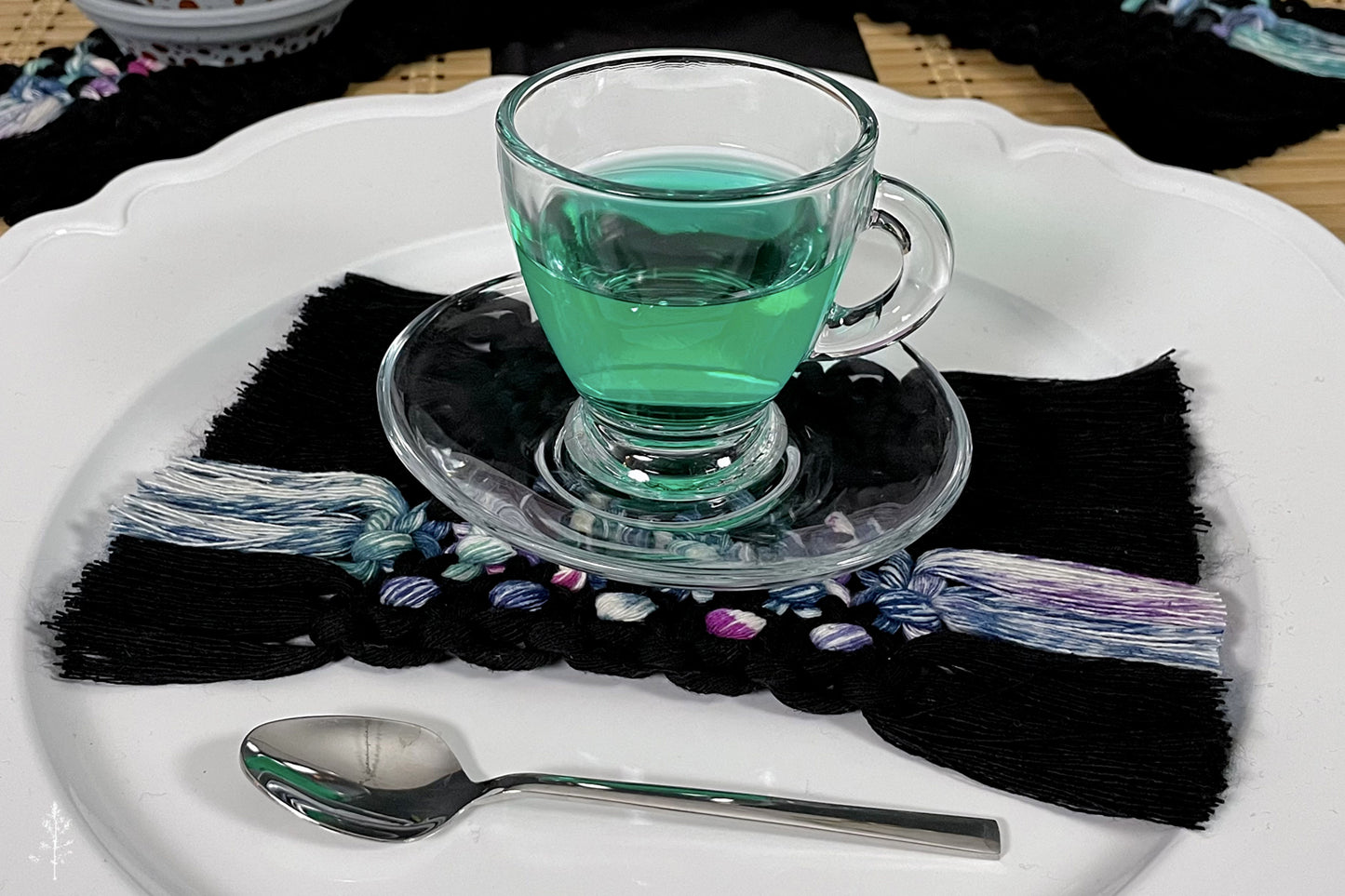 Black and tie dye macrame coaster. Green liquid in a glass on a decorative plate with a spoon